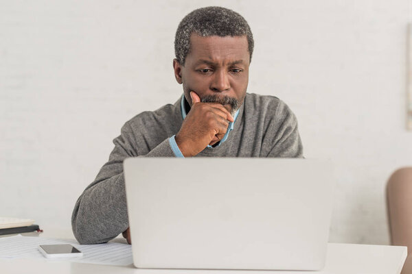 senior, thoughtful african american man looking at laptop while sitting at table