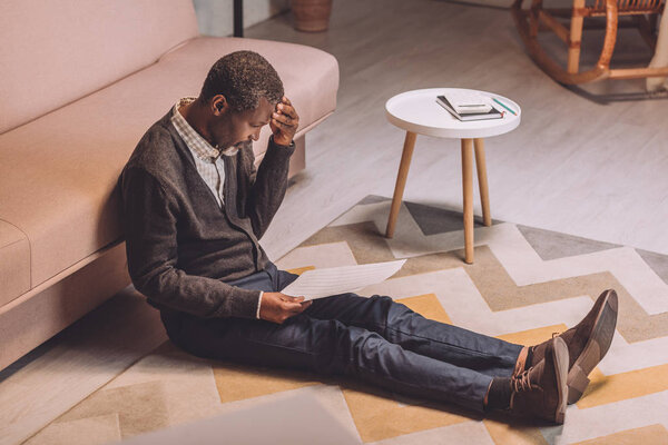 upset african american man sitting on floor and holding utility bill near round table with caulculator