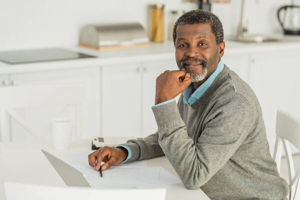 smiling african american man looking at camera while sitting at table near laptop