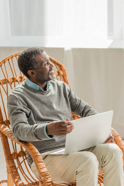 cheerful african american man sitting in wicker chair with notebook and looking away