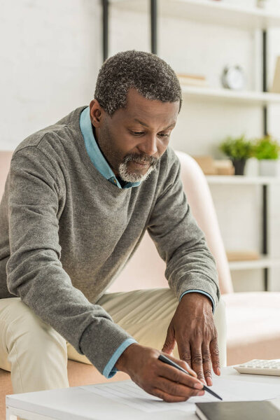 attentive african american man writing in utility bill while sitting on sofa