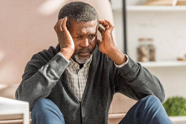upset african american man sitting on floor and holding hands near head while suffering from migraine