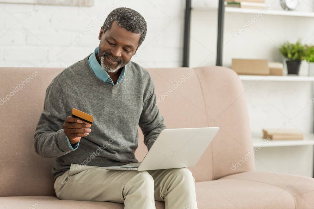 senior african american man sitting on sofa with laptop and holding credit card