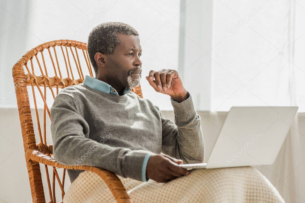 dreamy african american man sitting in wicker chair with laptop and looking away