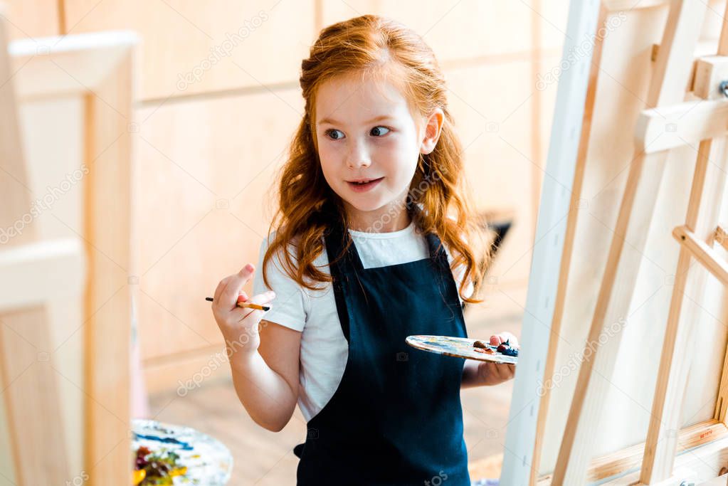 selective focus of cute redhead kid holding palette and looking away 