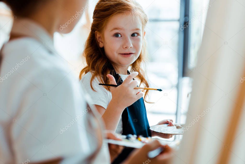 selective focus of cute redhead child looking at kid near easel 