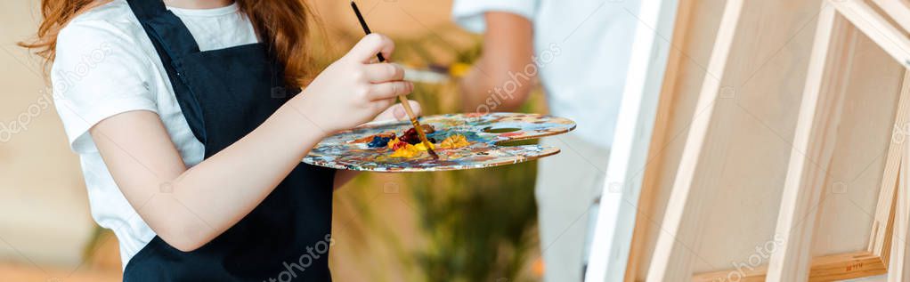panoramic shot of kid holding palette and paintbrush near easel 