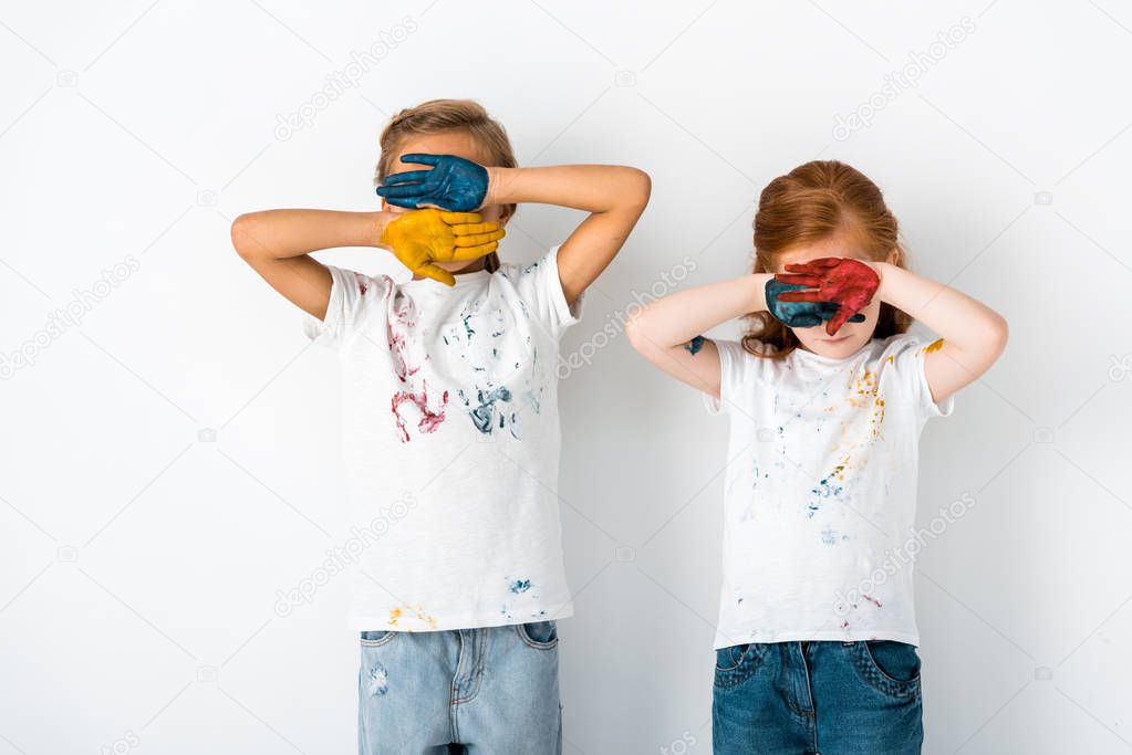 cute kids with paint on hands covering faces while standing on white 