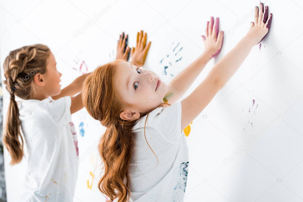 selective focus of happy redhead kid putting hand prints near child on white 