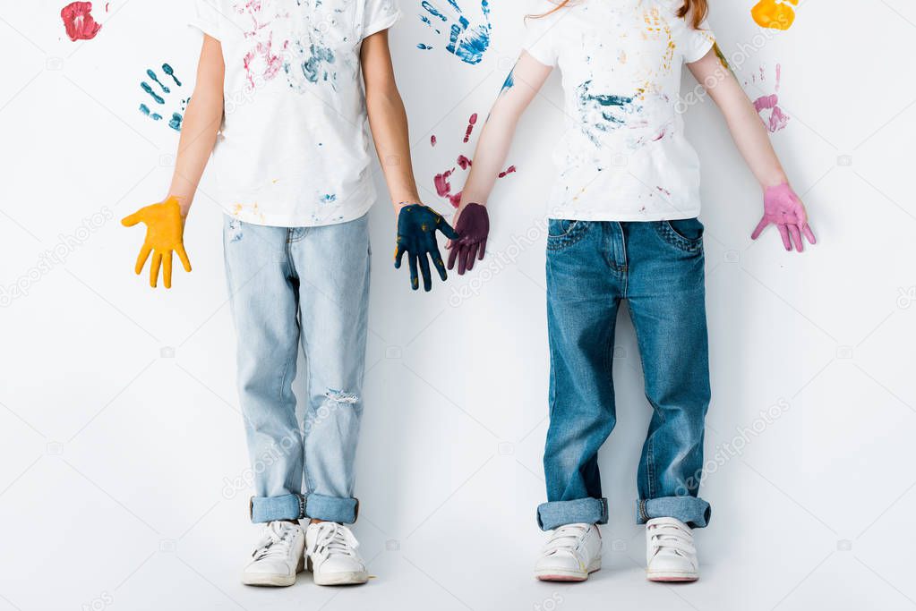 cropped view of kids in jeans with paint on hands standing near hand prints on white 