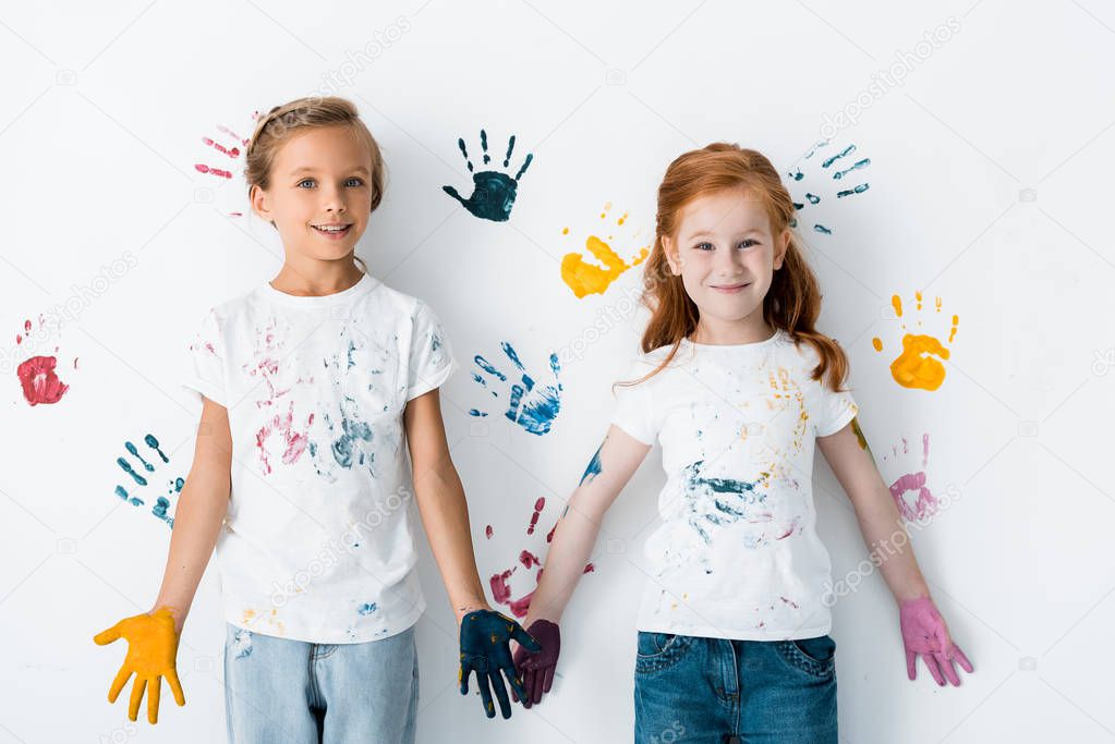 happy children with paint on hands standing near colorful hand prints on white 