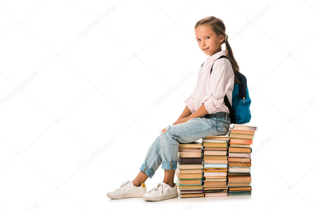 positive schoolkid sitting on books and looking at camera on white 