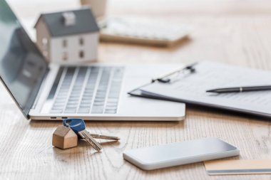 Bilgisayarın, akıllı telefonun, panonun ve ahşap masadaki ev modelinin yanındaki anahtarların seçici odak noktası