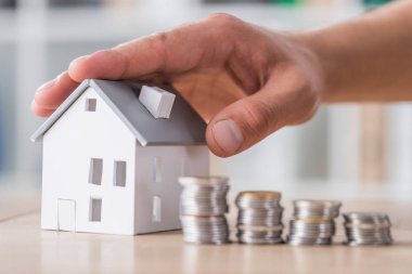 cropped view of businessman touching roof of house model near stacked coins clipart