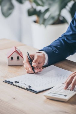 İş adamının hesap makinesi kullanması ve ev modelinin yakınındaki sözleşmede yazması.