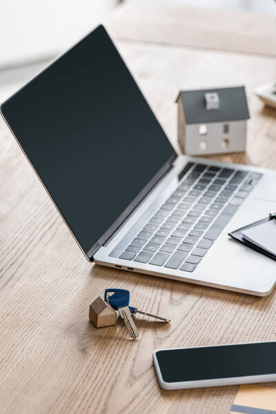 laptop and smartphone with blank screen, keys and house model on wooden table