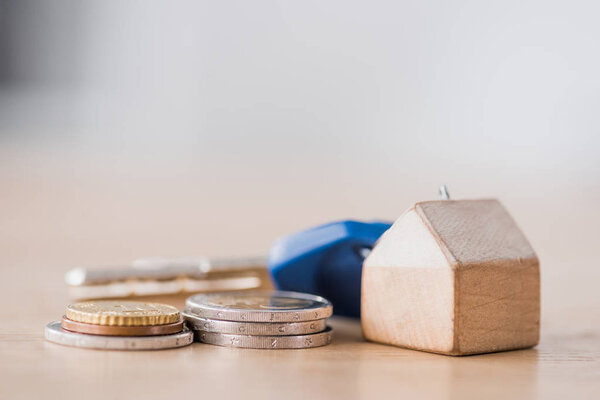 selective focus of toy house trinket with keys near golden and silver coins on wooden table