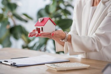 partial view of businesswoman holding house model near clipboard and calculator clipart