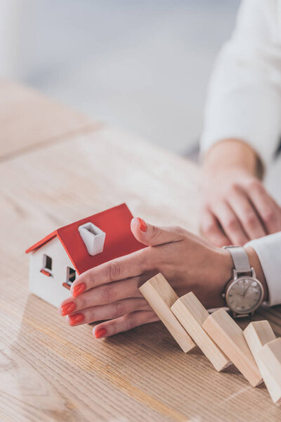 partial view of risk manager protecting house model from falling wooden blocks with hands