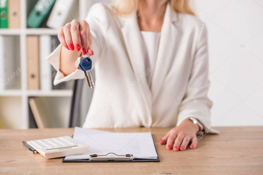 partial view of businesswoman at workplace holding keys and touching house model near clipboard and calculator
