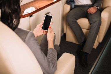 cropped view of businesswoman holding smartphone with blank screen near businessman in private jet  clipart