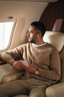 Sakallı adam özel jette müzik dinlerken elinde akıllı telefon tutuyor. 