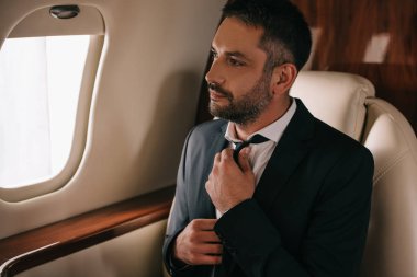 Sakallı iş adamı kravata dokunuyor ve uçakta oturuyor.