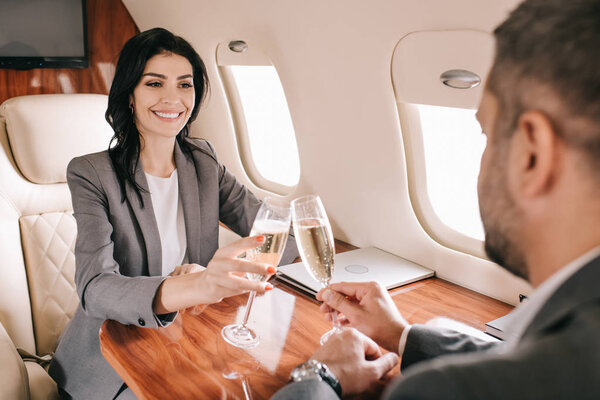 selective focus of cheerful businesswoman toasting champagne glasses with bearded businessman in private jet 