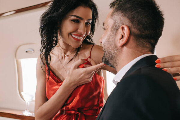 selective focus of cheerful woman in red dress touching face of bearded man in private jet 