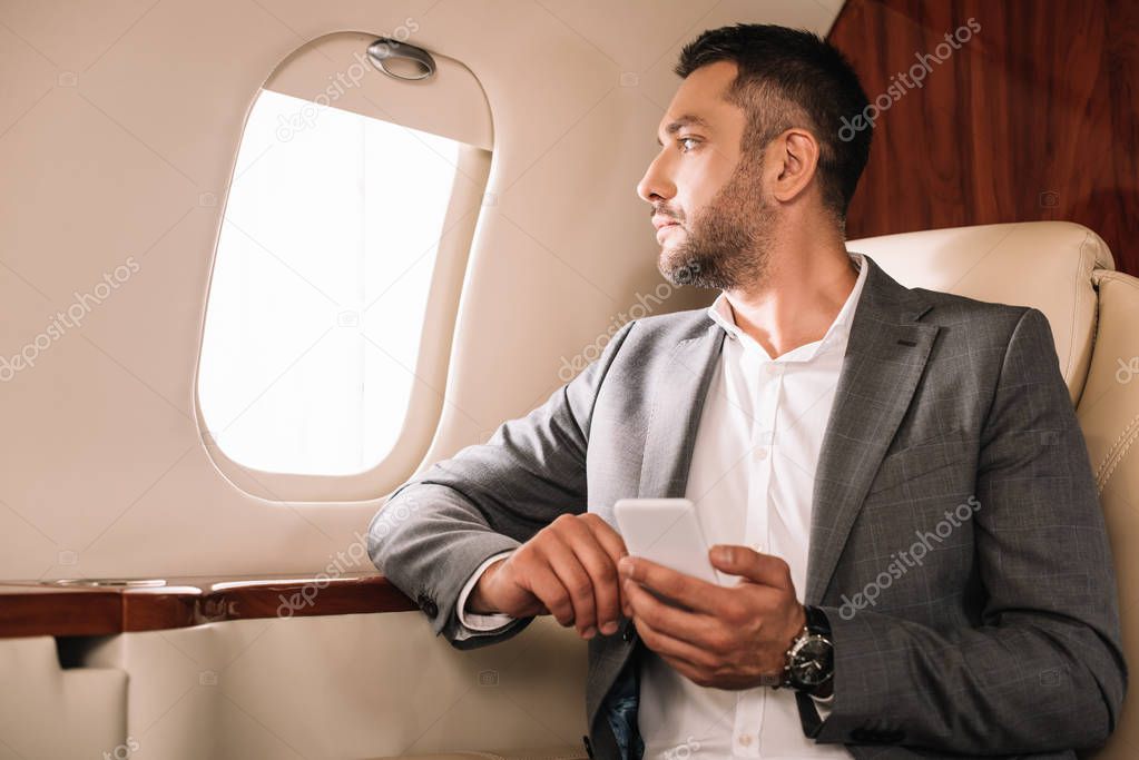 handsome businessman looking at airplane window while using smartphone in private jet 