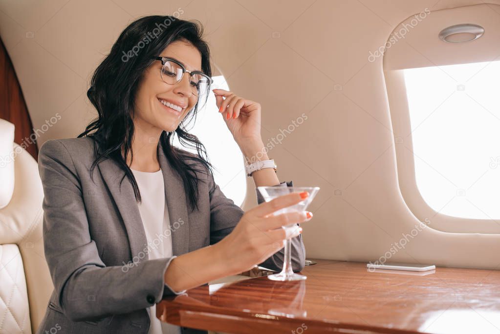 cheerful businesswoman in eye glasses holding martini glass in private jet 