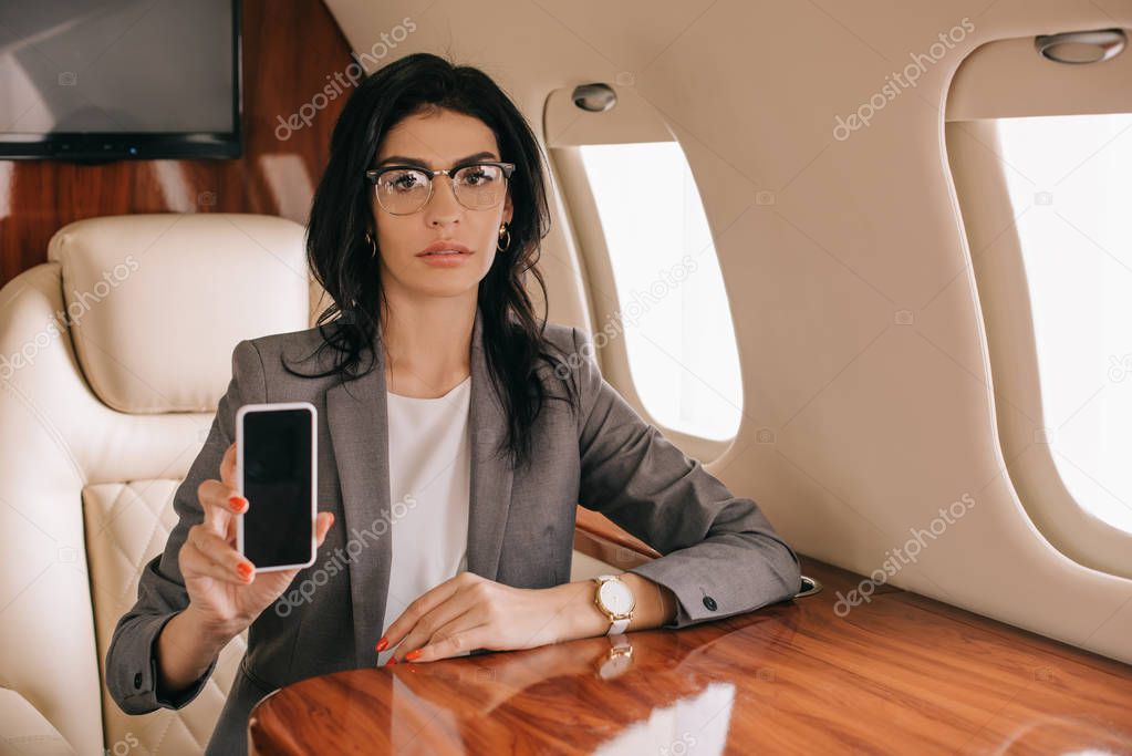 attractive businesswoman in glasses holding smartphone with blank screen in private jet 