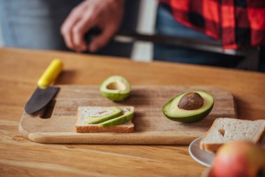 cropped view of man near chopping board with bread, knife and halves of fresh avocado clipart