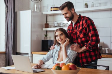 Bilgisayarın ve defterin yanındaki masada oturan gülümseyen kız arkadaşın omuzlarına dokunan yakışıklı adam.