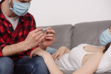 cropped view of man in medical mask looking at thermometer near sick girlfriend lying on sofa clipart