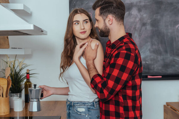 young man touching shoulder of smiling girlfriend making coffee in geyser coffee maker