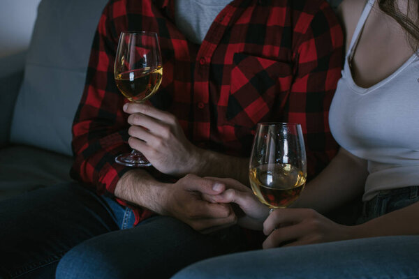 cropped view of couple holding hands while sitting on sofa with glasses of white wine