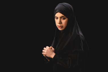 Side view of upset muslim woman in hijab with clenched hands isolated on black  clipart