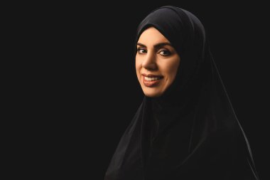 Beautiful muslim woman in hijab smiling at camera isolated on black  clipart