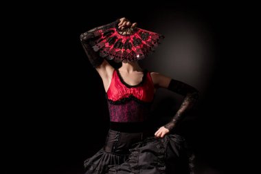 young flamenco dancer in dress covering face with fan on black clipart