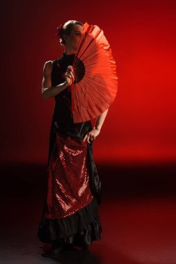 elegant flamenco dancer in dress covering face with fan while dancing on red  clipart