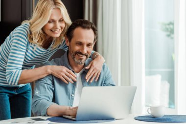 Mutfakta dizüstü bilgisayar kullanarak kocasını kucaklayan gülümseyen kadının seçici odağı. 
