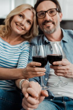 Gülümseyen olgun bir çiftin el ele tutuşup evde şarap içmesinin seçici odağı. 