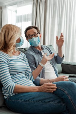 Tıbbi maskeli bir kadının seçici odağı kocanın elini kanepeye koyduğunu gösteriyor. 
