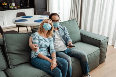 Kanepede film izlerken tıbbi maske takan olgun bir adam. 