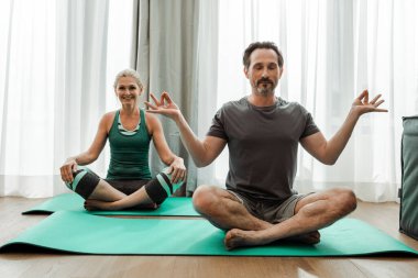 Oturma odasında gülümseyen karısının yanında meditasyon yapan olgun bir adam.