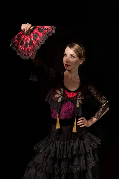 pretty flamenco dancer standing with hand on hip and looking at fan isolated on black