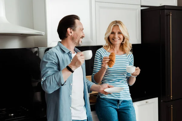 笑顔女性保持クロワッサン近く夫とともにコーヒーカップでキッチン — ストック写真
