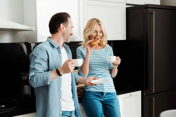 笑顔男保持コーヒーカップと見ます妻食べるクロワッサンでキッチン — ストック写真