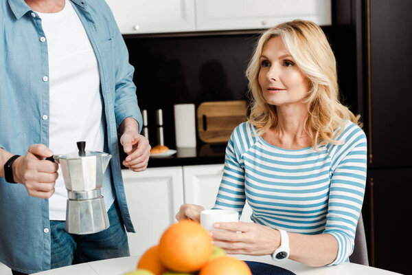 Selective focus of beautiful woman holding cup of coffee near man holding geyser coffee maker in kitchen 